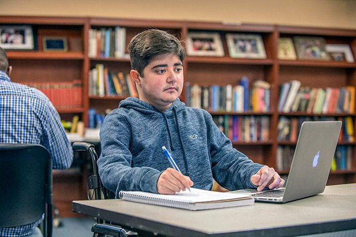 Student in a wheelchair looking at a laptop and taking notes in a notebook in a library.